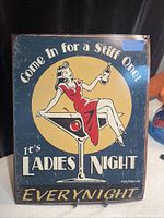 Photo of a rectangular vintage metal sign with a pin-up woman illustration and text about Ladies Night.