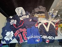 Photo showing multiple sports apparel items laid out including Turtle Leafs hoodies and t-shirts, New York sports jersey, Blue Jays shirt, Canada jacket, and Hamilton Tiger Cats hoodie.