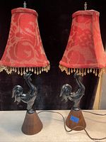 Pair of antique cast iron rooster table lamps with wood bases and red fabric lampshades featuring gold bead fringe. Electrical cords and plugs visible, both lamps standing upright.