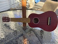 Full view of the 21-inch red ukulele laying on glass table outdoors around a wooden post, showing wooden neck and white tuning pegs.