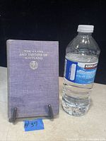 Full view of book standing upright next to water bottle for scale, showing purple cloth cover with gold title and emblem.