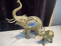 Photo showing side view of both brass elephant figurines, larger with raised trunk and tusks, smaller with raised trunk, placed on a white surface.