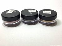 Three small clear jars with black lids of BareMinerals loose mineral eye color eyeshadows from top view showing lids with prices.