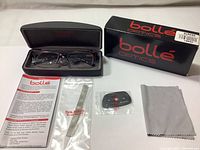 Eyeglasses in Bolle branded case with box, cleaning cloth, instructions, and accessories arranged on white background.