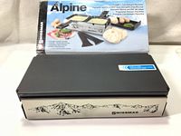 Closed Swissmar Alpine portable candlelight raclette in front of original box showing product branding and mountain design on stainless steel body.