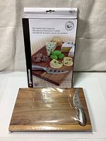 Mini acacia wood cutting board and stainless steel cheese cutting tool in front of the product box showing branding and product image.