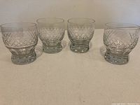 Four clear crystal lowball glasses showing crosshatch diamond pattern on sides, thick weighted bases, placed in a row on a light-colored surface against a plain background.