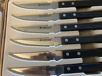 Set of six steak knives in foam tray, showing full set with black handles and serrated blades