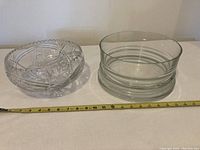 Two bowls on a white surface, one crystal bowl with cut pattern and one clear glass bowl with ring pattern. Tape measure visible showing size.