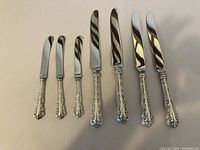Photo of 4 dinner knives and 3 butter knives with detailed scrollwork pattern on handles.