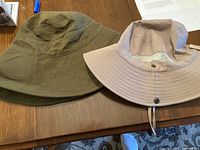 Two fabric wide brim hats on a wooden table, one khaki with mesh lining and chin strap, one olive green plain hat