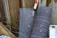 Two rolls of hexagonal chicken wire leaning against a wooden fence, showing a largely intact wire mesh with some outdoor dirt and leaves caught in the wire.