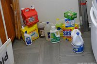 Photo showing assortment of large liquid cleaning supply jugs and boxes on floor against wall