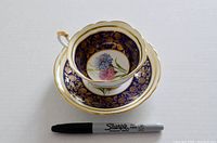Tea cup placed inside matching saucer with black Sharpie marker for scale, showing detailed floral center and blue gold patterned border.