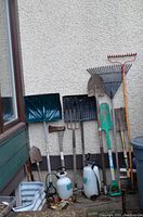Photo showing a variety of outdoor garden tools including shovels, rakes, shears, sprayers, and an axe leaning against an exterior building wall.