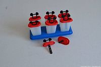 Front view of six popsicle molds with red lids topped with black Mickey Mouse ears handles on a blue stand; three lids removed showing the mold shape; one mold handle lying flat.