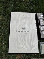 Plastic picture frame with backing showing brand 'Hobby Lobby' and size '24 x 36 inches' printed on it, placed on grass.