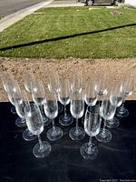 19 clear glass champagne flutes arranged in rows on a black surface outdoors.