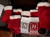 Six Christmas stockings in various sizes on a dark leather surface. Two patterned stockings with the letters 'N' and 'H' prominently shown. Other stockings are solid red with white cuffs or fur trim.