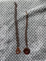Photo showing two necklaces on a textured gray fabric: one with a triangular orange pendant and thicker chain, the other with a circular dark brown hollow pendant and thinner chain.