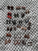 Photo showing a collection of 23 pairs of costume jewelry earrings arranged on a textured gray surface. Various styles include studs, hoops, dangles, in colors such as brown, pink, red, black, silver, and orange. Many earrings feature faux pearl or enamel accents.