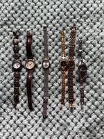 Photo showing six wristwatches arranged in a row on a grey textured surface, displaying different band styles and face shapes.