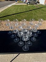 15 clear glass long stem candleholders arranged closely together outdoors on a black surface.