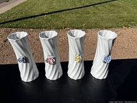 Photo of four tall white porcelain vases arranged side by side outdoors on a black surface with green grass background. Each vase has a spiral fluted texture and an applied flower in purple, pink, yellow, and blue respectively.