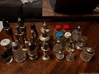 Multiple vintage salt and pepper shakers arranged on wooden table, featuring a variety of glass and metal designs and different sized caps including colored plastic tops.