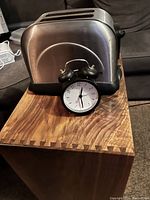 Photo showing side view of Bella stainless steel toaster and black analog alarm clock placed on wooden surface.