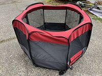 Red and black collapsible hexagonal playpen set up outdoors showing mesh ventilation panels and structural frame.