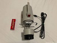 Side and top angle view of the vintage Elmo slide projector with power cord coiled and a box containing a spare lamp placed beside it.