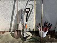 Full view of garden tools arranged against a wall, showing metal spade, Fiskars weeder, garden rake, white pail with pruning shears and nozzles, and roll of weed barrier plastic