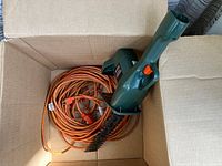 Photo shows green Black and Decker hedge trimmer with orange extension cord inside cardboard box.