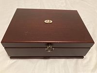 Closed dark wooden jewelry box with hinged lid and gold-tone emblem.