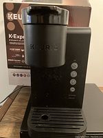 Front view of black Keurig K-Express Essentials coffee maker with control buttons and drip tray visible. Original product box in the background.
