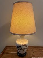 Full view of Chinese-painted ceramic table lamp with beige fabric lampshade illuminated showing bird and floral design.