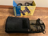 Two Bushnell PowerView 10x25 binoculars, one outside of its case with strap, other inside cases, both accompanied by original packaging boxes.