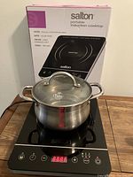 Salton portable induction cooktop with Palm Restaurant pot and glass lid in front. Cooktop black with controls and display, box in background.