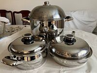 Three pieces of polished stainless steel cookware: two lidded saucepans with black handles and one large pot with dome lid and black handles.