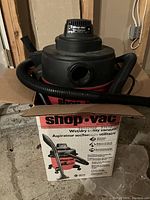 Shop Vac wet/dry vacuum seen inside partially closed original box; red and black body with label on motor unit and attached hose.