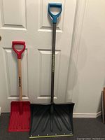 Two snow shovels standing upright against a white door, showing overall length and color of shovels and handles.