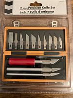 Front view of one 17-piece precision knife set showing multiple blades, blade holder and three knife handles in an orange plastic case inside sealed package.