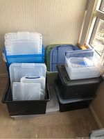 Photo of assorted plastic storage bins including clear, blue stackable drawers, black and green large containers with lids in corner of room