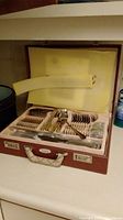 Opened case showing neatly arranged flatware pieces including forks, knives, spoons, serving utensils, and napkin rings inside cream lining.