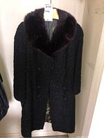 Full frontal image of a black Persian lamb fur coat with a dark brown fur collar hanging on a wall.