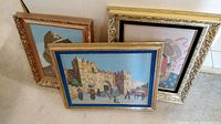 Three framed cross stitch pieces leaning against a wall, one with a castle scene and blue border, another with a woman in traditional dress in an ornate gold frame, and a third partially visible.