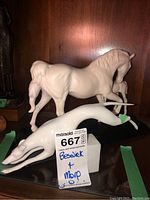 Two porcelain figurines displayed side by side; white running greyhound in front and standing horse behind with visible damage on the dog figurine's front leg.