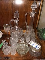 Full lot of glassware and crystal items including decanters, glasses, dishes, and vase arranged on a wooden shelf.