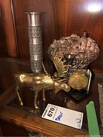 Photo showing brass moose figurine, pewter vase, Jerusalem cityscape plaque, and decorative medallion on a shelf.